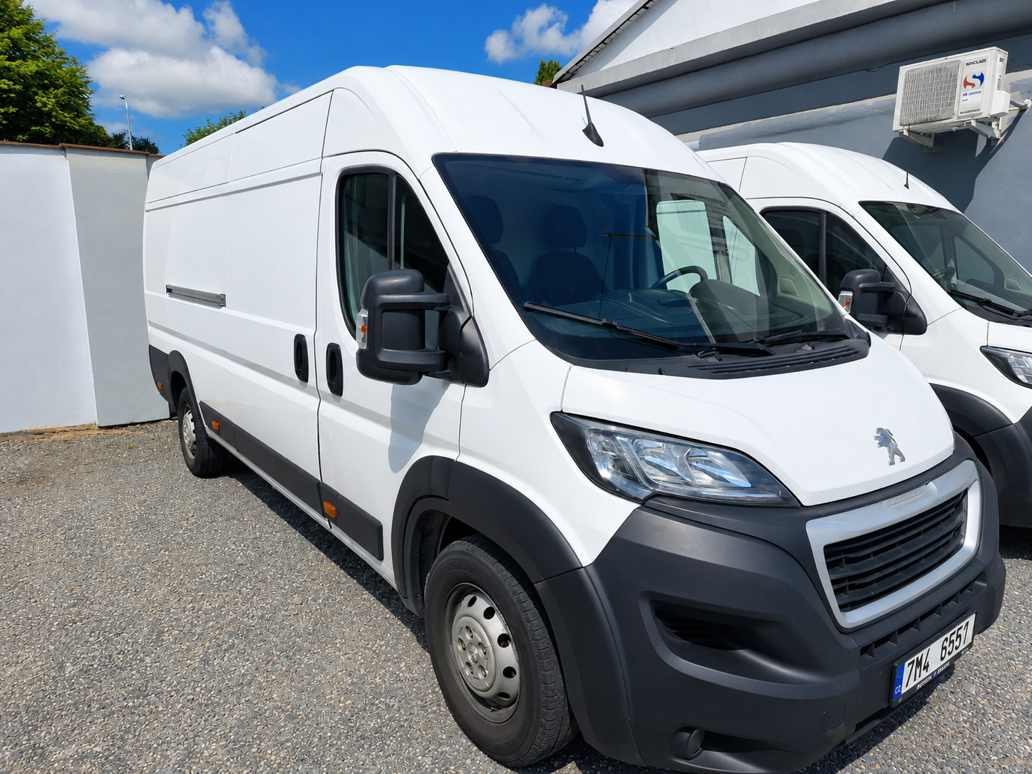 Bílý dodávkový vůz Peugeot s klimatizací na střeše, zaparkovaný u zdi, pod modrou oblohou.